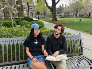 two students sitting on a bench on campus looking at a book together
